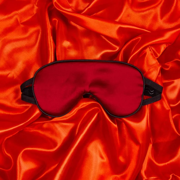Dark red silk sleep mask lie on bright red silken background. Top view. Square orientation