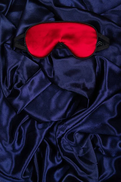 Bright red silk sleep mask lie on dark blue silken background. Vertical banner with negative space for copy placement