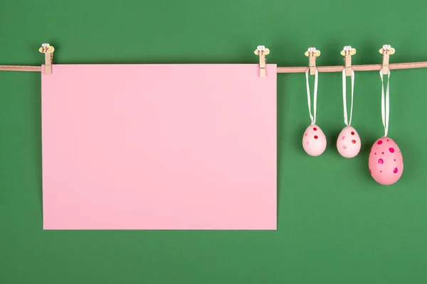 Blank pink sheet of paper and three pink Easter eggs hang at rope on green background. Easter card with room for your own message