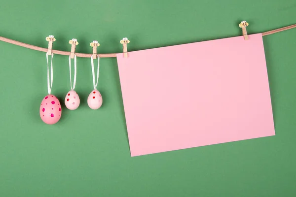 Blank pink sheet of paper and three pink Easter eggs hang at rope on green background. Easter greeting card design with empty space for text