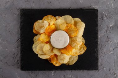 Gri alçı masa üzerinde servis panosu üzerinde kremalı beyaz soslu patates cipsi yığını manzarası. Lezzetli Cipsler ve Dip