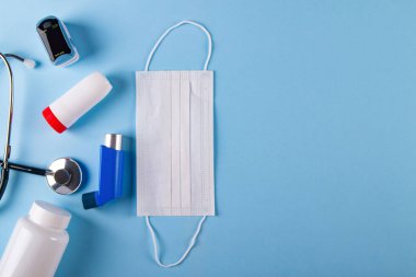 Tıbbi maske, stetoskop, oksimetre, ilaç şişesi ve mavi arka planda fotokopi alanı olan astım spreyi. Astımın yol açtığı solunum güçlüğü kavramı