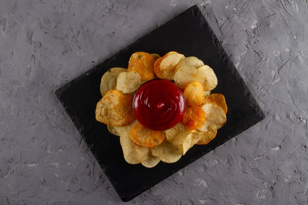 Top view of heap of potato chips with red dipping sauce on black slate serving board on grey plaster table. Elegant Entertaining Platter