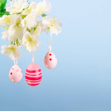 Dala asılı pembe yumurtaların hoş bir Paskalya gösterisi. Mavi arka planda çiçeklerle dolup taşıyor.