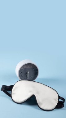 Beyaz ipek uyku maskesi ve alarm saati pastel mavi arka planda izole edilmiş. Dünya uyku günü konsepti