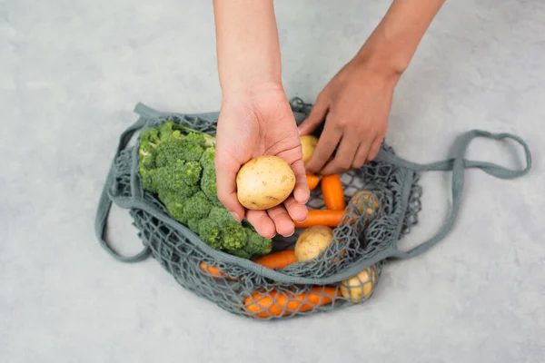 Hands taking potato from the mesh bag with vegetables, shopping grocery, healthy food ingredients, broccoli and carrots