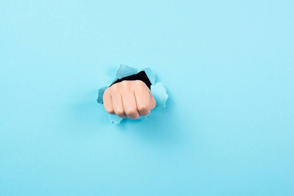 Fist brakes through torn ripped paper, punshing hole into blue background, conflict, stress and struggle concept