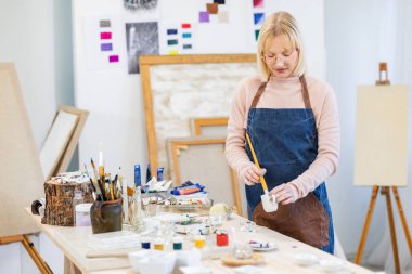 Young artist is mixing paint in her atelier, she is standing hear the working table with brush and paints