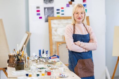 Young artist is posing in her painting studio, she is standing near her working table