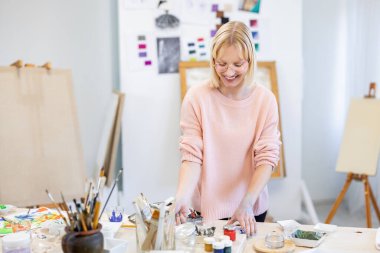 Genç kadın resim stüdyosunda. Çalışma masasının önündeki kameraya gülümsüyor.