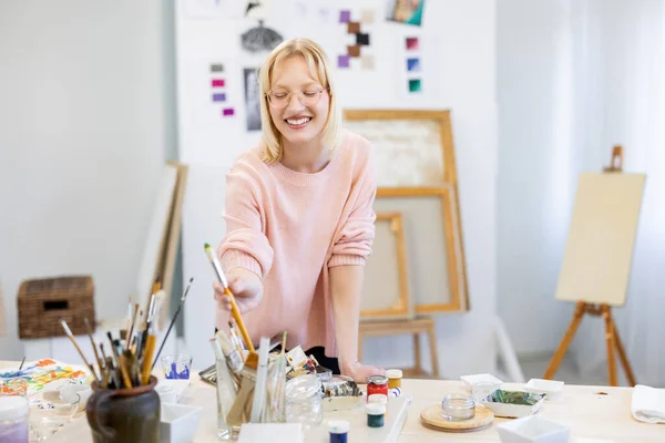 Genç Kadın Resim Stüdyosunda Çalışma Masasının Önündeki Kameraya Gülümsüyor — Stok fotoğraf