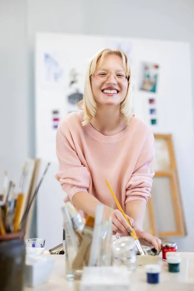 Genç Kadın Resim Stüdyosunda Çalışma Masasının Önündeki Kameraya Gülümsüyor — Stok fotoğraf