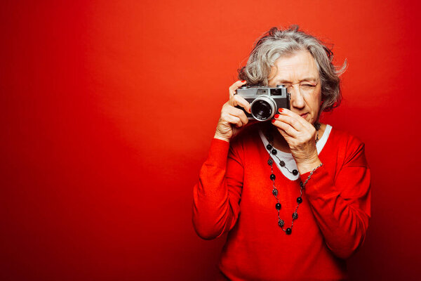 Старшая женщина в красной одежде, фотографирующая со старой кинокамерой, на красном фоне