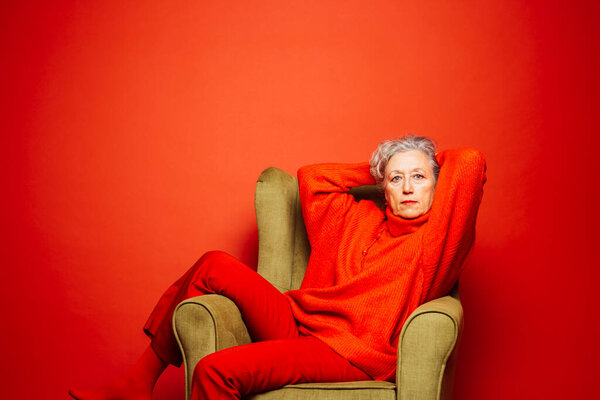 Senior woman wearing red clothes and sitting on a green armchair over a red background