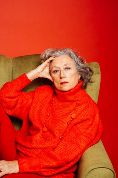 stock image Senior woman wearing red clothes and sitting on a green armchair over a red background