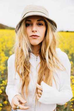Sarışın, güzel, neşeli bir kadının portresi, sarı kolza tohumu çiçekleri arasında bir şapka takıyor.