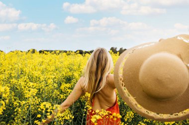 Kırmızı elbiseli, sarı kolza tohumu çiçekleri arasında yürüyen genç bir sarışının arka görüntüsü.