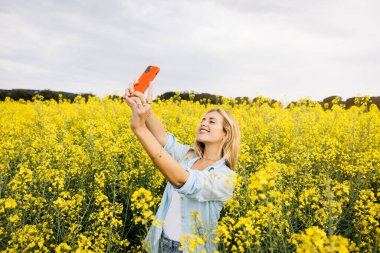 Güzel sarışın, neşeli kadın, kırmızı telefonuyla selfie çekiyor, sarı kolza çiçeği tarlasının ortasında.