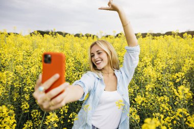 Güzel sarışın, neşeli kadın, kırmızı telefonuyla selfie çekiyor, sarı kolza çiçeği tarlasının ortasında.