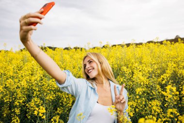 Güzel sarışın, neşeli kadın, kırmızı telefonuyla selfie çekiyor, sarı kolza çiçeği tarlasının ortasında.