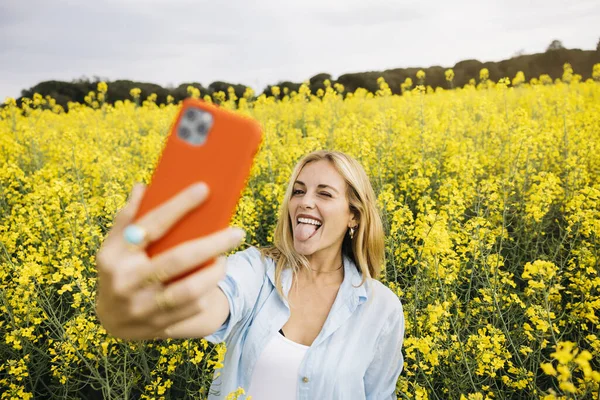 Güzel sarışın, neşeli kadın, kırmızı telefonuyla selfie çekiyor, sarı kolza çiçeği tarlasının ortasında.