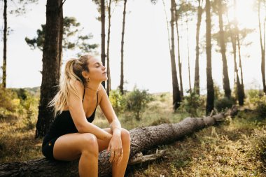 Genç bayan koşucu bir ağaç gövdesinde dinlenmek için duruyor. Spor salonundaki kadın ormanda antrenmana ara veriyor..
