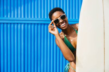 Bir sörfçünün portresi ayakta, mayolu mavi bir arka planda, yanında sörf tahtasıyla. Yaz tatilinde plaj aktiviteleri yapan mutlu bir kadın..