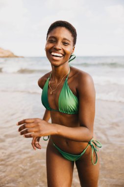 Sahilde deniz suyundan kaçan bikinili, neşeli, gülümseyen siyah kadın.