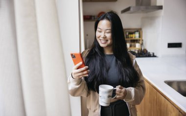 Genç, güzel bir kadın elinde kupayla mutfakta pencerenin yanında kırmızı bir telefon kullanıyor. Gülümseyen kadın evde kahve molası veriyor..