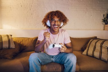 Beyaz kulaklıklı genç bir adam oturma odasındaki bir kanepede video oyunları oynuyor. Mutlu genç adam zaferi kutluyor.