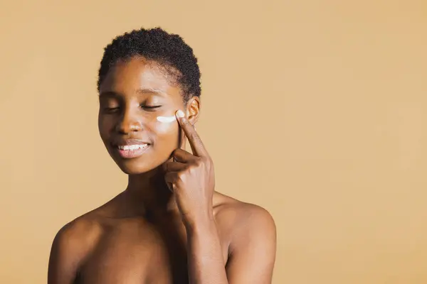 Young black smiling woman with eyes closed applying moisturising cream on her face. Young woman treating her facial skin with a beauty product.