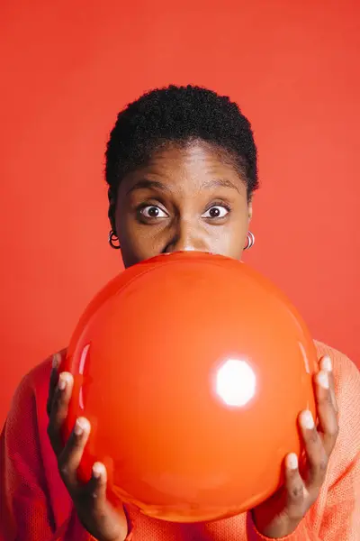 Happy young woman blowing a red balloon in front of red background in a studio. Woman with short hair inflating a red balloon.