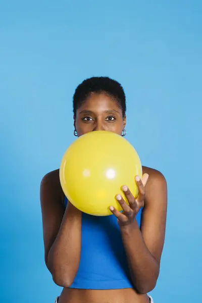 Young woman blowing a yellow balloon in front of blue background in a studio. Woman with short hair inflating a yellow balloon.
