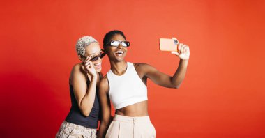 Güneş gözlüğü takan ve stüdyoda kırmızı arka planda selfie çeken neşeli genç bir kadının portresi. Stüdyoda iki oyuncu kadın birlikte..