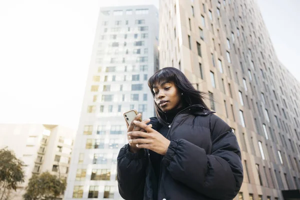 Young beautiful black woman wearing black clothes typing an online message on her phone while walking on the street of a city.