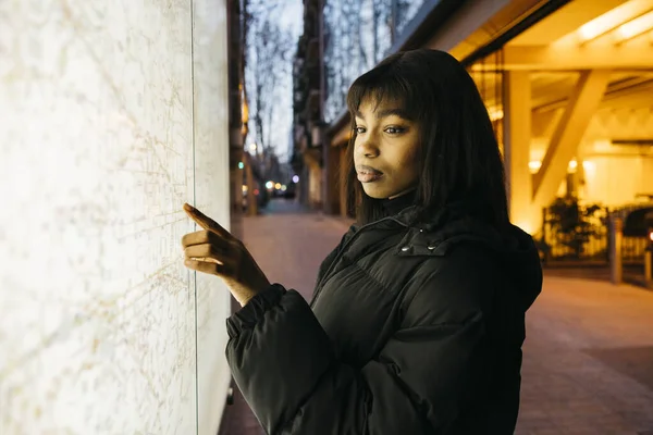 Young black woman looking at a city map on a bus stop in the street.