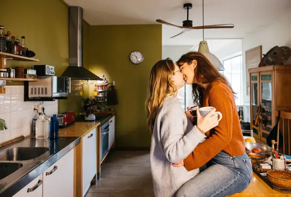 Young female couple kissing while sharing a cup of tea in a in their home kitchen. Lesbian couple enjoying a moment together, sipping and tea and kissing in the comfort of their home kitchen.