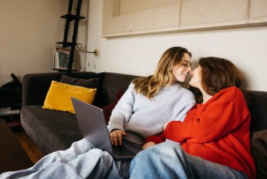 Kanepede oturmuş laptoplarıyla birbirlerini öpen lezbiyen çift. Genç eşcinsel çift samimi ve samimi bir atmosferde dizüstü bilgisayar kullanıyor..