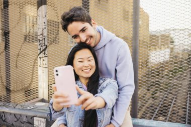 Şehir demiryolunun üzerinde oturmuş gülümseyen ve telefonuyla selfie çeken bir çift. Irklar arası bir çift takılıyor ve selfie çekiyor..