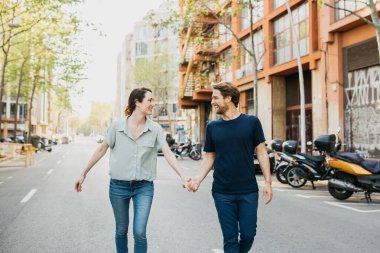 Çiftler el ele yürür bir şehir caddesinde, gülümseyerek ve birbirlerine bakarak, güneşli bir günün tadını çıkararak. 