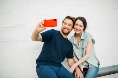 Çift birlikte oturuyor, gülümsüyor ve kırmızı bir telefonla beyaz bir duvara karşı selfie çekiyor. İfadeleri mutluluk ve yakınlığı gösteriyor..