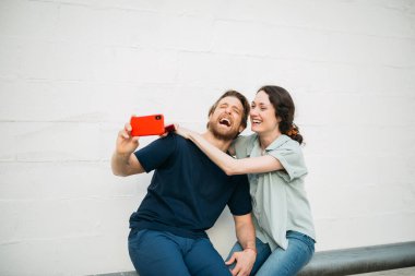 Çift birlikte oturuyor, gülümsüyor ve kırmızı bir telefonla beyaz bir duvara karşı selfie çekiyor. İfadeleri mutluluk ve yakınlığı gösteriyor..