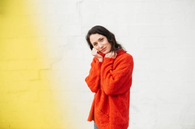 Kırmızı kazaklı bir kadının portresi beyaz tuğladan bir duvara karşı duruyor, kazağının yakasını yüzüne yakın tutuyor ve nazikçe gülümsüyor..