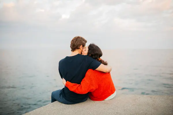 stock image Couple sitting together on a pier, embracing and looking out over the calm sea under a cloudy sky, sharing a peaceful moment.