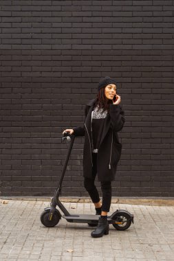 Rasta bukleli genç bir kadın elektrikli scooter 'da kırmızı bir akıllı telefonla konuşurken gülümsüyor, siyah tuğla bir duvarın önünde duruyor..