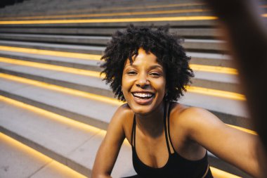 Aydınlatılmış basamaklarda oturan neşeli genç bir kadın bir şehir ortamında idman yaptıktan sonra selfie çekiyor..