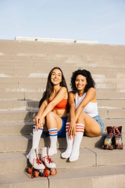 Merdivenlerde oturmuş, gülümseyen ve sohbet eden iki arkadaşın portresi. Her biri renkli çoraplı patenler giyiyor ve paten yapmaya ara vermenin keyfini çıkarıyor..