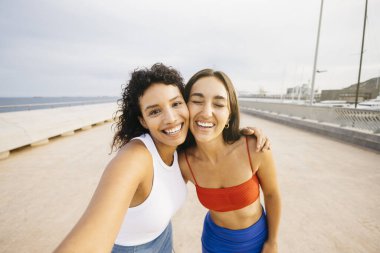 İki bayan arkadaş bir deniz kenarında selfie çekiyor, sarılıyor ve gülümsüyor..