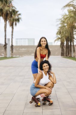 İki arkadaş fotoğraf için poz veriyor, biri paten ile çömelmiş, diğeri ise arkasında duruyor, hem gülümsüyor hem de şehir parkında eğleniyorlar..