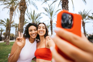 İki arkadaş, arka planda palmiye ağaçları olan, gülümseyen ve sarılan kırmızı bir telefonla selfie çekiyorlar..